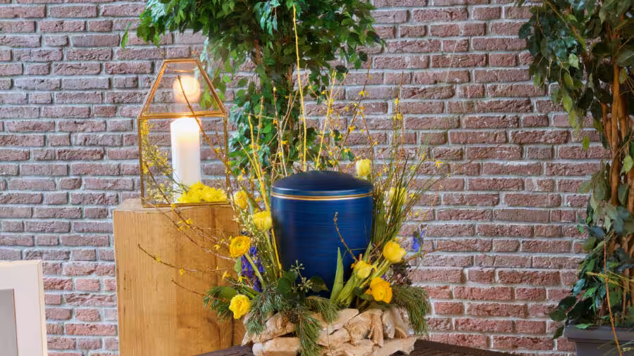 Urne mit Blumenarrangement vor Ziegelwand.
